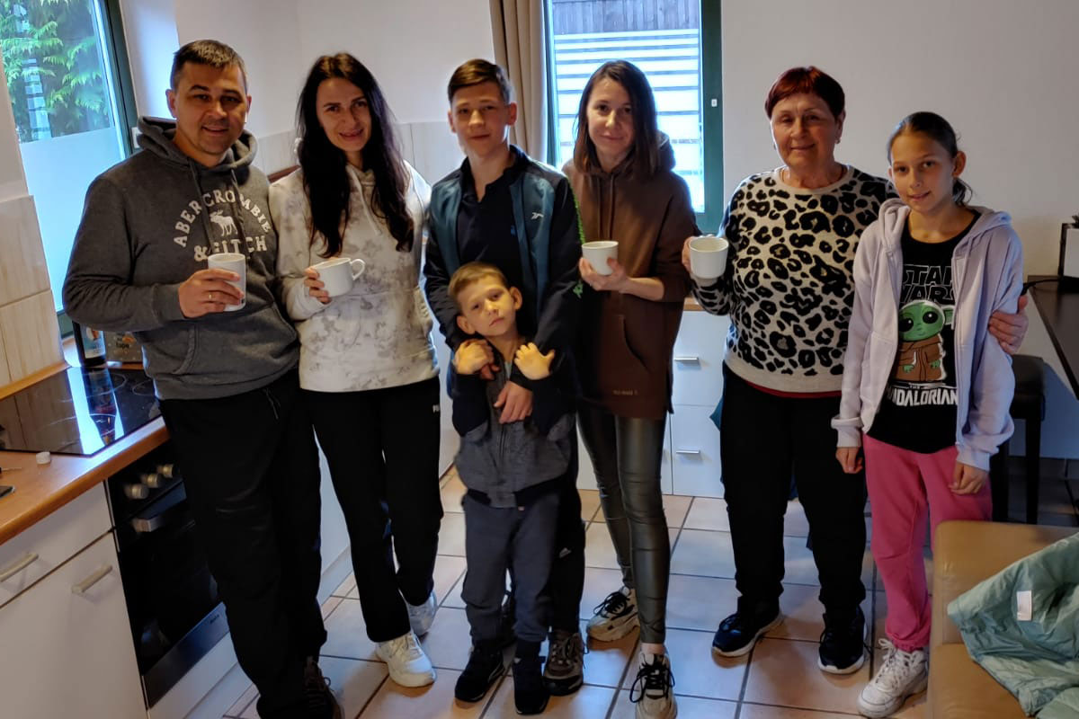 Vater, Mutter, vier Kinder und die Großmutter stehen lächelnd in ihrer großen, hellen Küche.